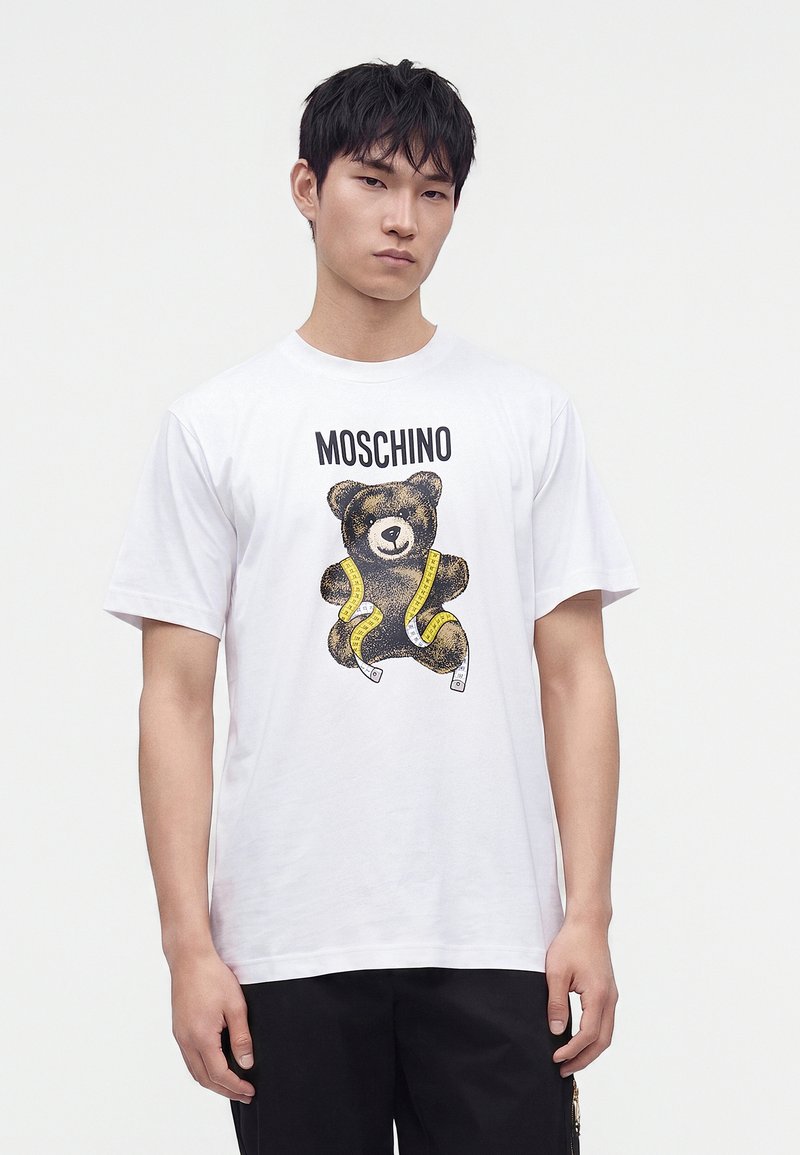Νέος άνδρας που φορά ένα λευκό t-shirt Moschino με σχέδιο αρκουδάκι και κίτρινη μεζούρα, στέκεται μπροστά σε απλό φόντο.