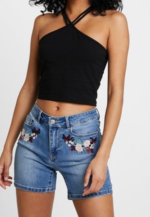 Vrouw in een zwarte halter crop top en hooggerande blauwe denim shorts met bloemenborduursel op de voorzakken.