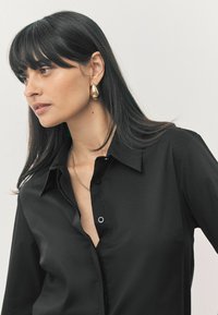 Zwarte blouse met kraag, met een zachte textuur en een iets ontspannen pasvorm, gedragen met een gouden druppeloorbel en ketting. 