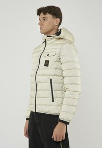 Giacca in crema imbottita con cappuccio, zip frontale, dettagli neri, tasca sul petto e polsini elasticizzati, che presenta un motivo trapuntato e materiale resistente.