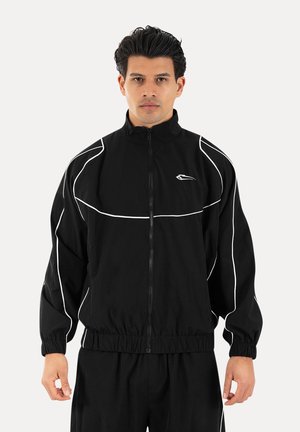 Hombre con chaqueta negra con cremallera y detalles blancos y pantalones negros a juego, de pie frente a un fondo claro y liso.