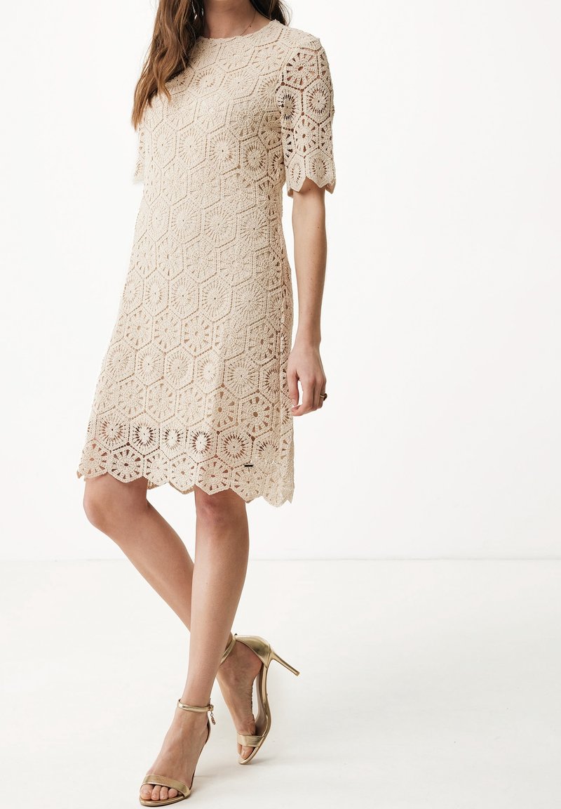 Robe en dentelle à motif hexagonal, couleur beige, manches courtes, ourlet festonné, portée avec des sandales à talons hauts dorées sur un fond clair.