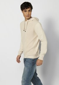 Hoodie beige clair en tissu tricoté texturé, avec un cordon de serrage, des manches longues et un ourlet côtelé. Porté avec un jean bleu.