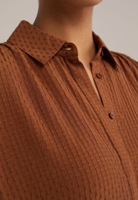 Camicia button-up marrone con un motivo testurizzato, caratterizzata da un colletto e strisce verticali. Si notano piccoli bottoni e un materiale morbido e leggero.