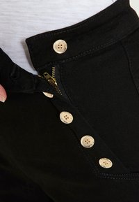 Pantalones negros con cierre de cremallera, cinco grandes botones en tono dorado y una tela texturizada cerca de la cintura con detalles de costura en contraste.