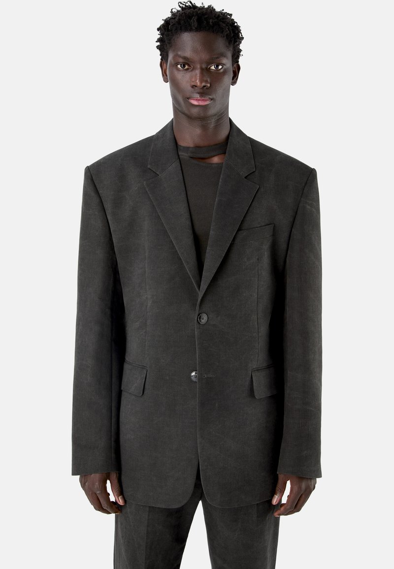 Młody mężczyzna ubrany w oversize'ową ciemnoszarą marynarkę na czarną bluzkę, stojący na jasnym, jednolitym tle.