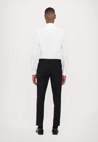 Weißes Langarmhemd mit erhöhtem Kragen und schwarze Hose. Die Hose ist tailliert mit schmaler Passform und hat Gesäßtaschen.