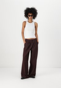 Cămașă albă fără mâneci și pantaloni evazați în nuanța burgundă. Modelul poartă ochelari de soare negri și pantofi negri cu accente argintii. Textura materialului este netedă.