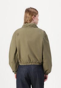 Giacca corta verde oliva con colletto alto, vita elastica e maniche a palloncino casual. Sul retro è presente un piccolo logo.