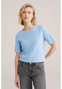 Vaaleansininen, teksturoitu neulottu lyhythihainen pusero, jossa on pyöreä pääntie ja geometrinen kuviointi. Yhdistetty korkeavyötäröisiin harmaisiin farkkuihin.