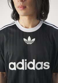 Fekete sportmez fehér "adidas" felirattal, bordázott textúrával és három fehér csíkkal a vállakon. Klasszikus logó a felirat fölött.