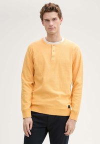 Suéter de manga larga y cuello redondo en algodón amarillo claro, con tapeta abotonada y puños y dobladillo acanalados. Etiqueta negra en la esquina inferior izquierda.