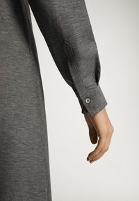 Bras portant une chemise grise texturée à manches longues avec un poignet boutonné, main détendue pendante sur un fond uni.