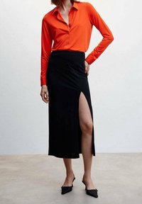 Chemise rouge à manches longues rentrée dans une jupe midi noire fendue jusqu'à la cuisse. La tenue est complétée par des chaussures noires à enfiler.