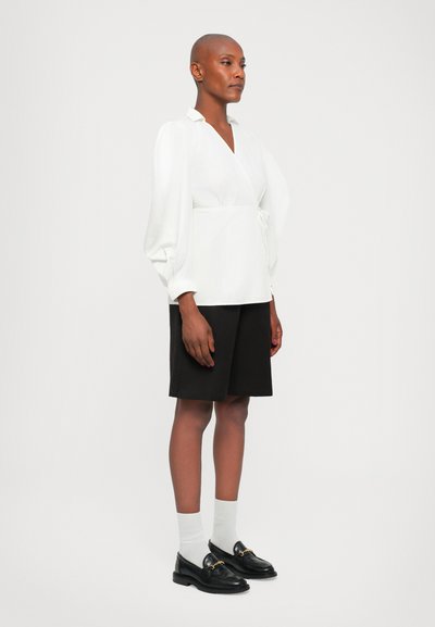 Witte wikkeltop met lange pofmouwen, gecombineerd met zwarte shorts. Model draagt zwarte loafers en witte crew-sokken. Eenvoudig en gestructureerd ontwerp.