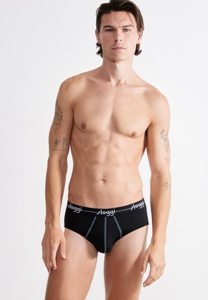 Jeune homme avec des tatouages sur les deux bras portant un slip noir Sloggi, debout devant un fond blanc uni.