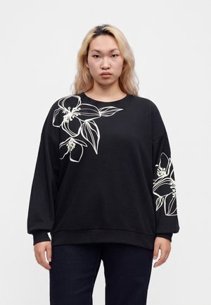 Ung person med langt blond hår iført sort oversized sweatshirt med store hvide blomsterdesigns på skulderen og ærmet.