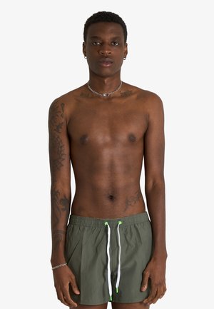 Giovane uomo con tatuaggi su braccia e petto, indossa pantaloncini da bagno verdi con coulisse bianche su uno sfondo semplice.