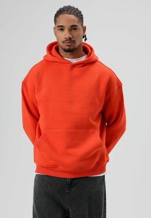 Oranje hooded sweatshirt met een voorzak en geribbelde zoom, gemaakt van zachte stof. Model draagt het met donkergrijze broek.