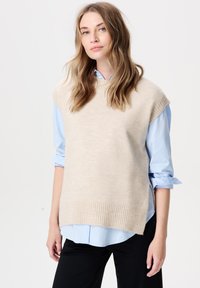 Beige gebreide mouwloze trui met een geribbelde zoom, gedragen over een lichtblauw overhemd met knopen, met opgerolde mouwen en neutrale zwarte broek.