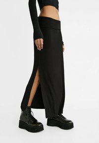 Schwarze, enganliegende Maxirock mit seitlichen Schlitzen, aus glattem Stoff gefertigt. Kombiniert mit massiven schwarzen Schnürstiefeln für einen kontrastierenden Look.