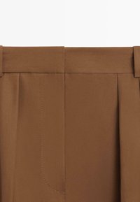 Zbliżenie na pas brązowych, dopasowanych spodni z szlufkami na pasek i subtelnymi przeszyciami.