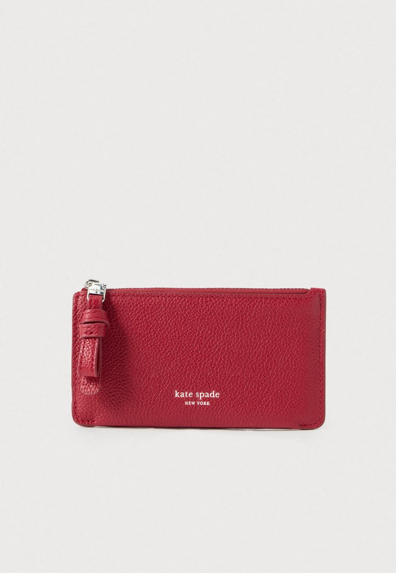 Červená kožená peňaženka s textúrovaným povrchom, zipsovým uzáverom a malým remienkom. Na prednej strane je vyrazený nápis "kate spade NEW YORK" v bielej farbe. Kompaktný dizajn.