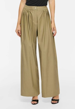 Femme portant un pantalon beige taille haute à jambes larges avec des plis devant, fermeture par boutons, assorti à des talons ouverts noirs et un haut noir.