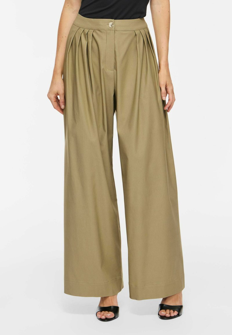 Femme portant un pantalon beige taille haute à jambes larges avec des plis devant, fermeture par boutons, assorti à des talons ouverts noirs et un haut noir.