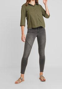 Chemise boutonnée vert olive, jean skinny gris à texture côtelée et panneaux d'accent, associée à des sandales beige avec un motif rayé.