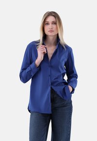Long-sleeve blue shirt with a pointed collar, button front, and relaxed fit. Paired with dark denim jeans. Smooth texture and solid color.