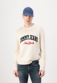 Pulover alb din tricot cu textul "TOMMY JEANS" și "New York" în culori bleumarin și roșu, asociat cu blugi denim închisi la culoare și o șapcă albastră.