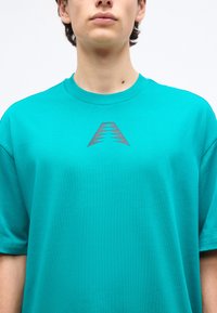 Personne portant un t-shirt turquoise à col rond avec un petit motif géométrique triangulaire sombre centré sur la poitrine.