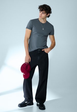 Giovane uomo con t-shirt grigia, pantaloni neri larghi e scarpe da ginnastica nere, che tiene un berretto rosso in una mano e guarda verso sinistra.