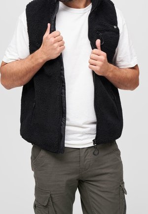 Mand iført hvid T-shirt, sort fleecevest og olivengrønne cargobukser med hænder, der griber fat i vestens kanter mod en ensfarvet baggrund.