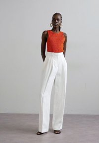 Κόκκινο υφασμάτινο αμάνικο μπλουζάκι σε συνδυασμό με λευκά παντελόνια ψηλής μέσης. Το μοντέλο φορά μεγάλα σκουλαρίκια κρίκους και καφέ σανδάλια με λουριά. Ουδέτερο φόντο.