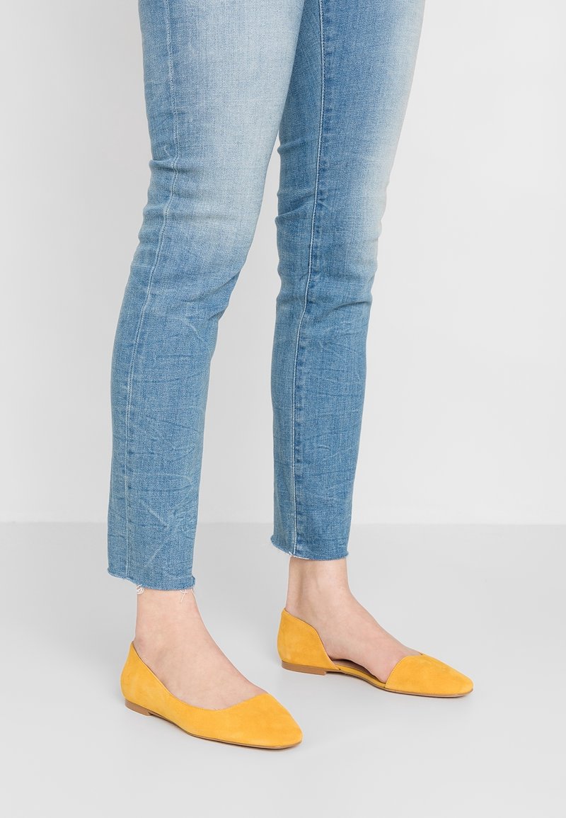 Jambes portant un jean skinny bleu clair et des ballerines pointues jaune moutarde, debout sur un sol gris clair devant un mur blanc.