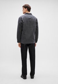 Férfi fekete függőleges csíkos zakó, gallérral, hosszú ujjal és texturált anyagból. Sötét, egyenes szárú nadrággal és cipővel párosítva.