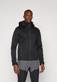 Giacca nera con chiusura a zip, cappuccio regolabile e due tasche laterali. Presenta una texture liscia e un design aderente, abbinata a pantaloni grigi.