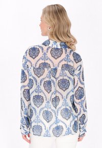 Durchsichtige Bluse in Creme mit floralem Muster in Blau und Dunkelgrau. Lange Ärmel mit Knopfmanschetten und leicht gewelltem Saum.