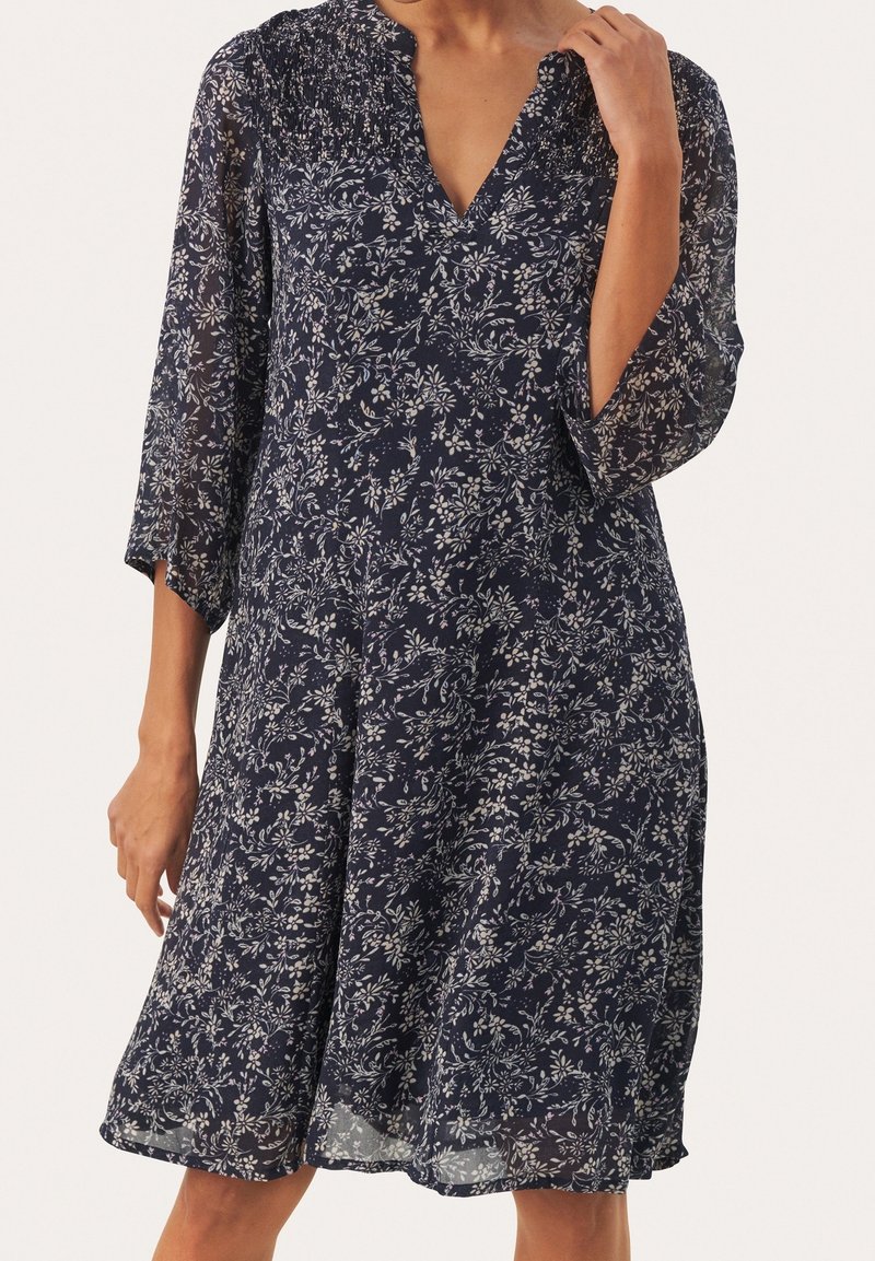 Femme portant une robe bleu marine longueur genou avec des motifs floraux blancs et des manches transparentes trois-quarts, debout devant un fond uni.