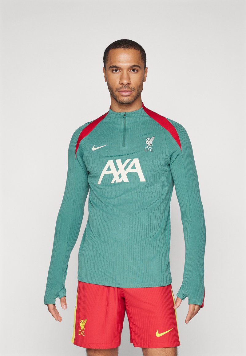T-shirt de sport vert à manches longues avec texture côtelée, accents rouges sur les épaules, logo blanc "AXA" et blason LFC, associé à un short rouge.