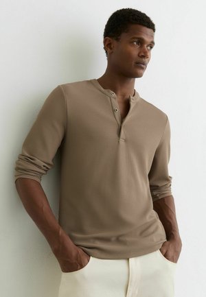 Uomo con maglia henley beige a maniche lunghe con maniche arrotolate e pantaloni bianchi, in piedi con le mani in tasca contro una parete bianca.
