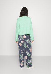 Osoba stojąca tyłem, ubrana w miętową bluzę, granatowe spodnie o szerokich nogawkach z kwiatowym wzorem oraz białe buty na jednolitym tle.