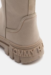 Beige saapas, joka on valmistettu teksturoidusta materiaalista ja jossa on selkeä ompelu. Saappaassa on paksu, uritettu pohja ja ''TOMMY HILFIGER'' -logo painettuna kantapään kohdalla.