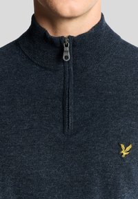 Maglione zip-up blu scuro con colletto a coste, caratterizzato da una trama grigia a fiocchi e un piccolo logo di un'aquila ricamato in giallo sul lato sinistro del petto.
