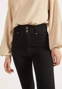 Jeans negros de cintura alta con una textura suave, que cuentan con dos botones metálicos y bolsillos laterales. La silueta es ajustada.