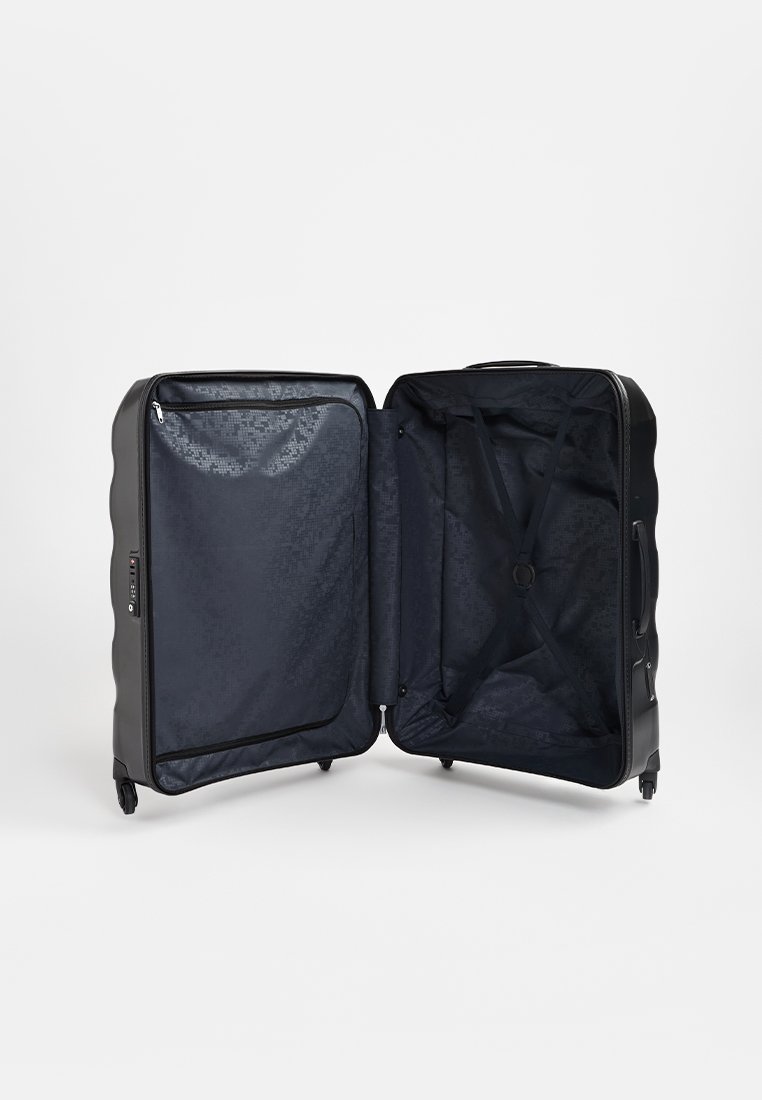 Valise noire rigide avec une surface texturée, double zips, sangles croisées intérieures et doublure à motifs. Ouverte et vide.
