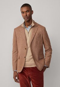 Brauner Fischgräten-Blazer aus strukturiertem Stoff, über einem beigen Reißverschluss-Pullover getragen. Ergänzt rote Cordhosen mit einem subtilen Muster.