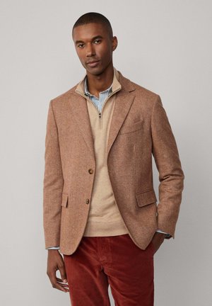 Blazer en chevrons marron en tissu texturé, superposé à un pull zippé beige. Complète un pantalon en velours côtelé rouge avec un motif subtil.